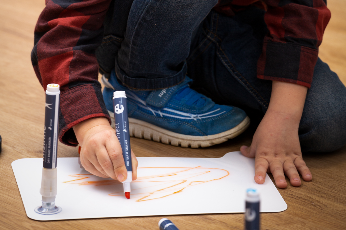 Ein Kind in blauen Turnschuhen und einem rotkarierten Hemd malt eifrig mit einem orangefarbenen Marker auf einem Whiteboard-Ideenbrett der imsinne GmbH, während andere Marker auf dem Holzboden um sie herum verstreut sind.